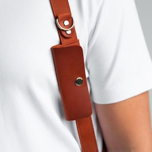 May include: Close-up of a brown leather shoulder strap with a small pouch and a gold-coloured metal clasp. The strap is attached to a white shirt. The leather has a smooth texture and is secured with stitching.