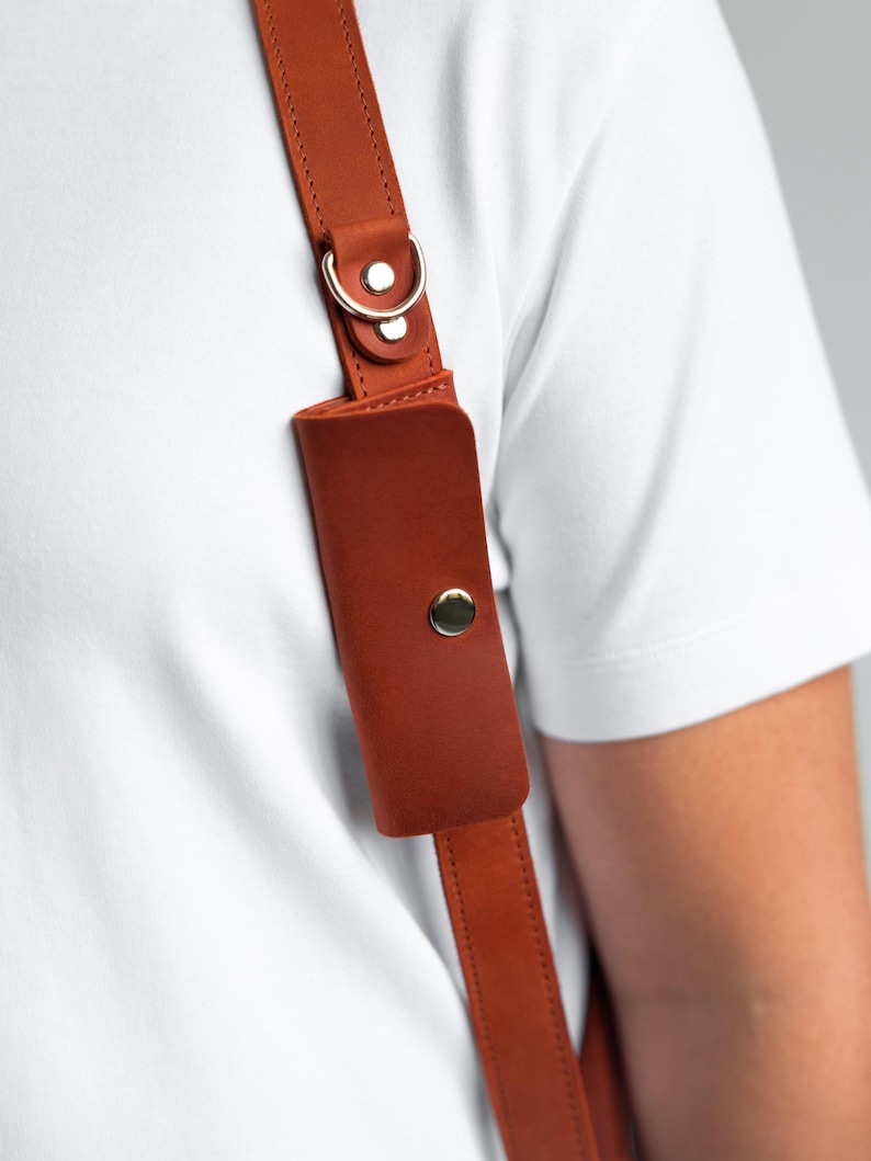 May include: Close-up of a brown leather shoulder strap with a small pouch and a gold-coloured metal clasp. The strap is attached to a white shirt. The leather has a smooth texture and is secured with stitching.