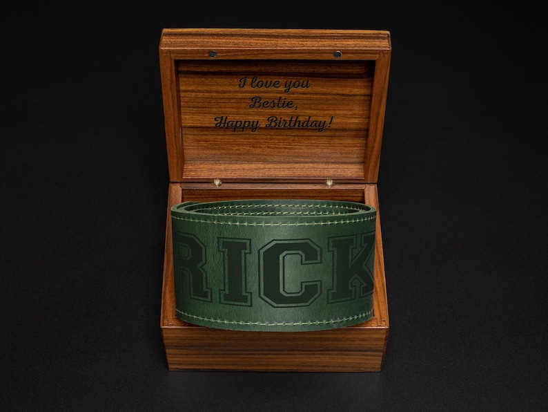 Puede incluir: Una caja de madera abierta, que revela un cintur&oacute;n de cuero verde con el nombre "RICK" en relieve. El interior de la caja tiene la inscripci&oacute;n "I love you Bestie, Happy Birthday!"