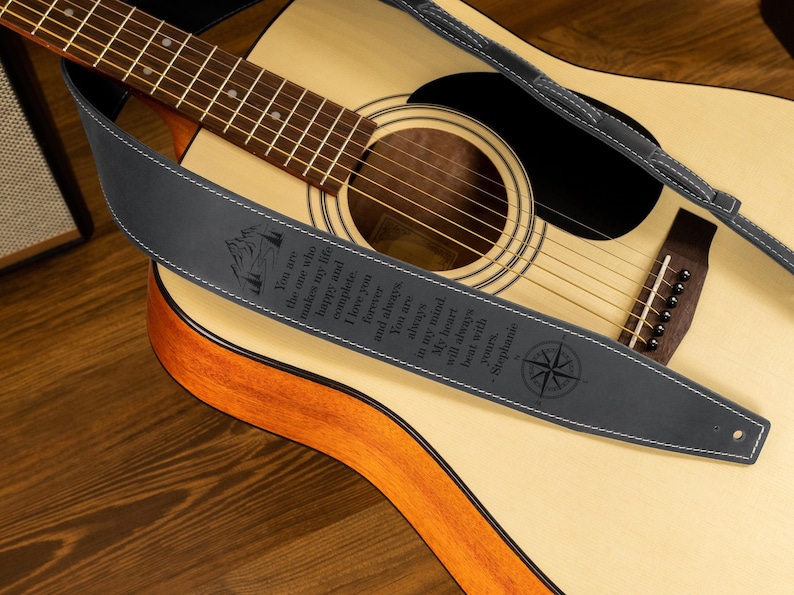 May include: Acoustic guitar with a dark gray leather guitar strap. The strap has white stitching and engraved text with a compass rose design. The guitar is light brown with a dark brown sound hole.