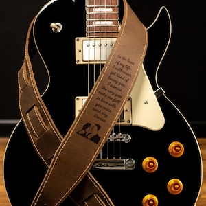 K&ouml;nnte beinhalten: Eine schwarze E-Gitarre mit einem braunen Ledergitarrengurt. Der Gurt hat wei&szlig;e N&auml;hte und einen eingravierten Text, der lautet "To the love of my life..." mit einer Silhouette eines sich k&uuml;ssenden Paares.