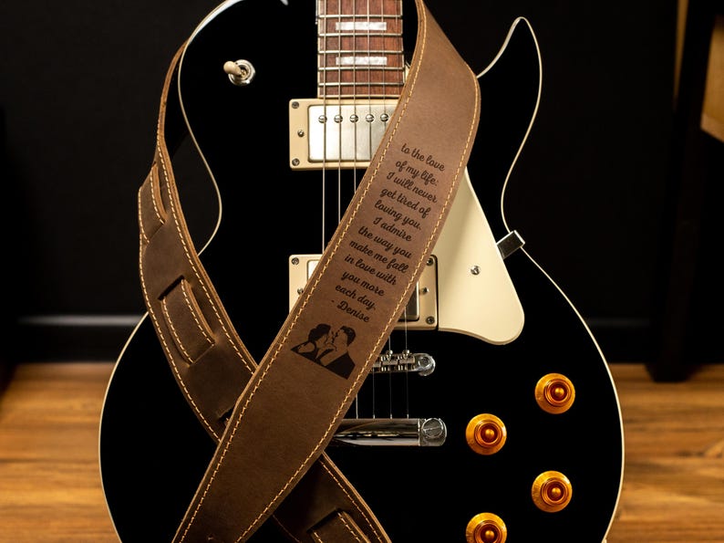 K&ouml;nnte beinhalten: Eine schwarze E-Gitarre mit einem braunen Ledergitarrengurt. Der Gurt hat wei&szlig;e N&auml;hte und einen eingravierten Text, der lautet "To the love of my life..." mit einer Silhouette eines sich k&uuml;ssenden Paares.