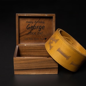Puede incluir: Una caja de madera con tapa con bisagras, grabada con "Happy Birthday, George ROCK ON!" y una correa de guitarra de cuero amarillo con las letras "KIL" en relieve. La caja y la correa est&aacute;n sobre una superficie negra.