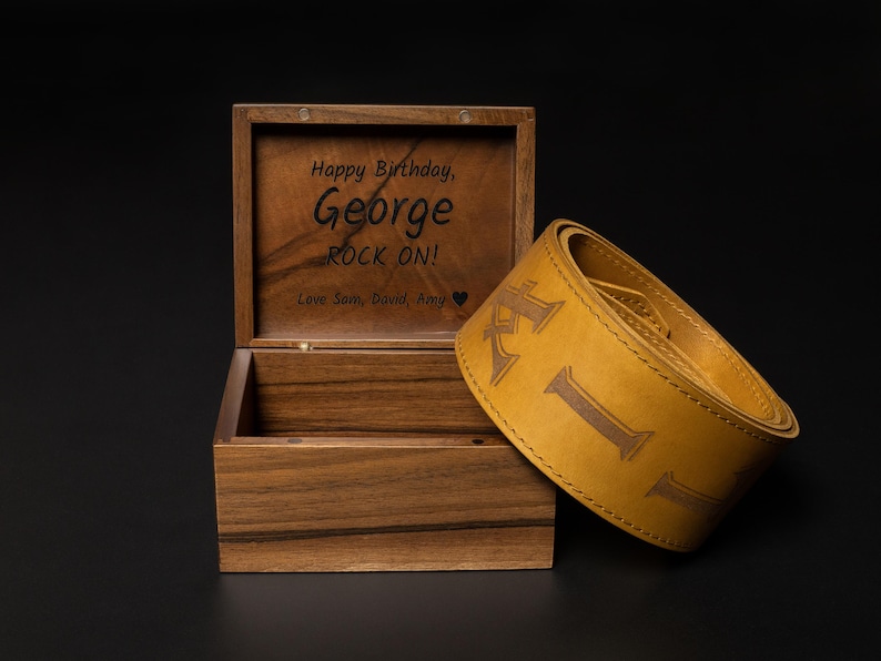 Puede incluir: Una caja de madera con tapa con bisagras, grabada con "Happy Birthday, George ROCK ON!" y una correa de guitarra de cuero amarillo con las letras "KIL" en relieve. La caja y la correa est&aacute;n sobre una superficie negra.