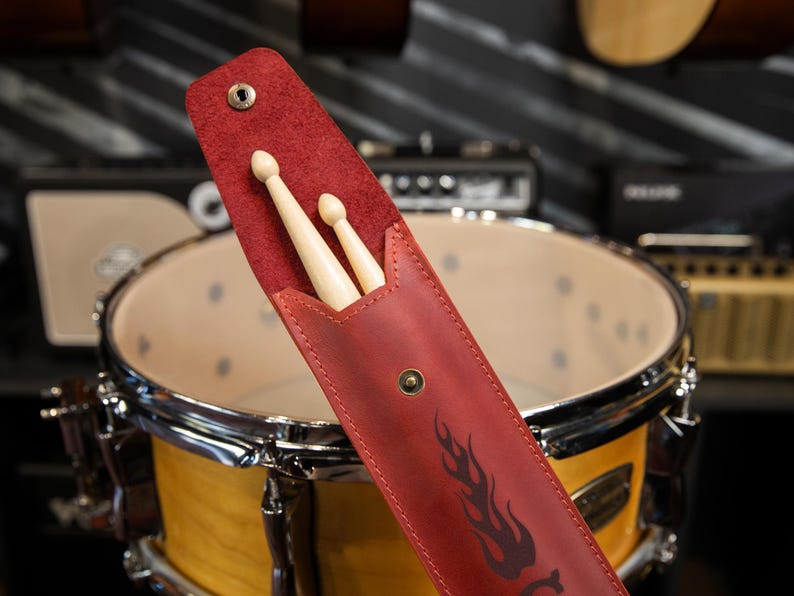 May include: A red leather drumstick case with a flame design, holding two wooden drumsticks. The case is open, revealing the sticks, and is positioned in front of a drum set.