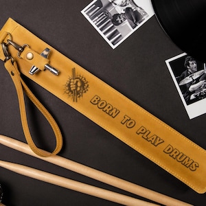 May include: A tan leather drumstick case with the text "BORN TO PLAY DRUMS" and a drum graphic. Includes a wrist strap and metal hardware. Two wooden drumsticks are also visible.