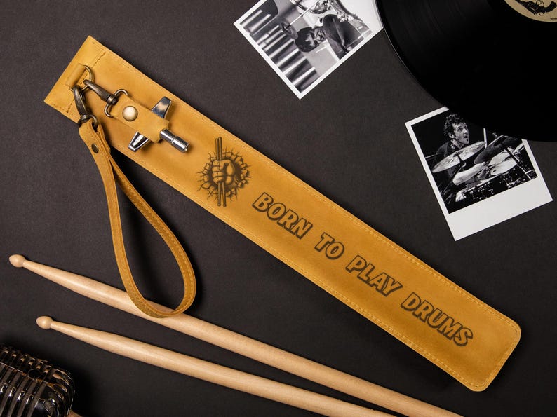 May include: A tan leather drumstick case with the text "BORN TO PLAY DRUMS" and a drum graphic. Includes a wrist strap and metal hardware. Two wooden drumsticks are also visible.