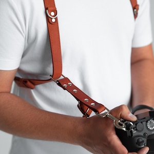 May include: A brown leather camera harness with silver metal fixings. The harness has adjustable straps and is designed to be worn over the shoulders. The camera is black and has a lens attached. The harness is designed for photographers.