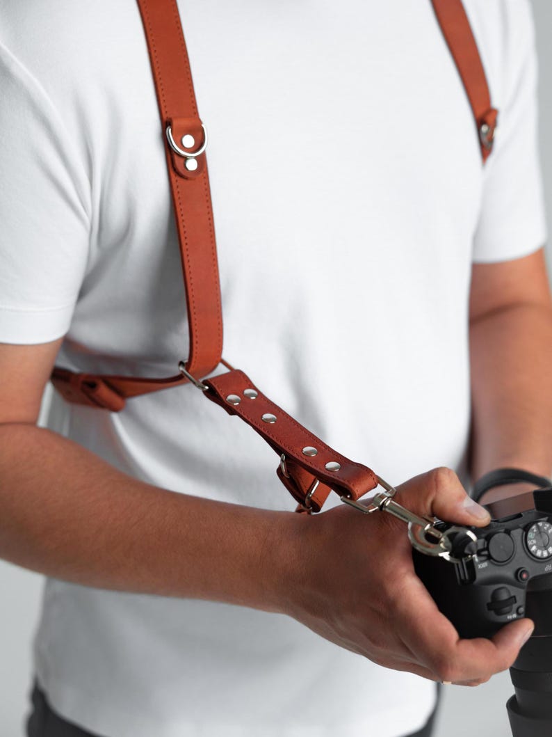 May include: A brown leather camera harness with silver metal fixings. The harness has adjustable straps and is designed to be worn over the shoulders. The camera is black and has a lens attached. The harness is designed for photographers.