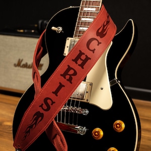 K&ouml;nnte beinhalten: Eine schwarze E-Gitarre mit einem roten Leder-Gitarrengurt. Der Gurt tr&auml;gt den Namen "SIRICH" und Flammendesigns. Im Hintergrund ist ein Marshall-Verst&auml;rker zu sehen.