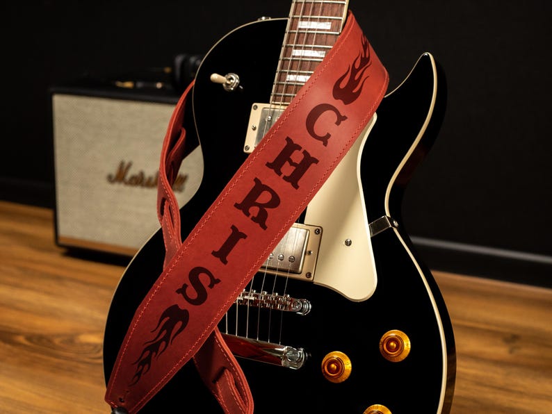 K&ouml;nnte beinhalten: Eine schwarze E-Gitarre mit einem roten Leder-Gitarrengurt. Der Gurt tr&auml;gt den Namen "SIRICH" und Flammendesigns. Im Hintergrund ist ein Marshall-Verst&auml;rker zu sehen.