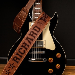Puede incluir: Una correa de guitarra de cuero marr&oacute;n con el nombre "RICHARD" grabado en letras grandes. La correa presenta un dise&ntilde;o de &aacute;guila y bandera, junto con detalles de estrellas. La correa est&aacute; colocada sobre una guitarra el&eacute;ctrica negra.