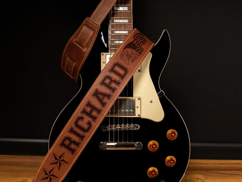 Puede incluir: Una correa de guitarra de cuero marr&oacute;n con el nombre "RICHARD" grabado en letras grandes. La correa presenta un dise&ntilde;o de &aacute;guila y bandera, junto con detalles de estrellas. La correa est&aacute; colocada sobre una guitarra el&eacute;ctrica negra.