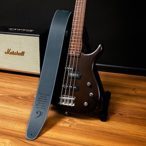 May include: A dark brown bass guitar with a blue leather strap. The strap has white stitching and a bass clef symbol. Text on the strap reads "Don't practice until you get it right, practice until you can't get it wrong."