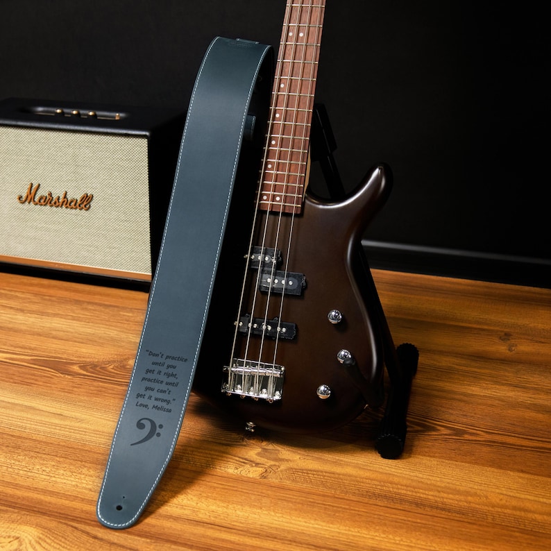 May include: A dark brown bass guitar with a blue leather strap. The strap has white stitching and a bass clef symbol. Text on the strap reads "Don't practice until you get it right, practice until you can't get it wrong."