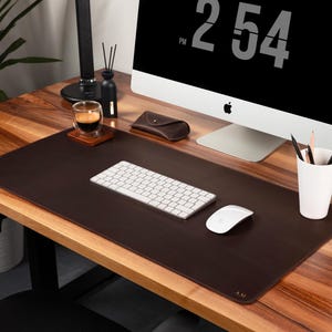 Op de afbeelding: Een donkerbruine leren bureauonderlegger met een wit toetsenbord en muis. Een computermonitor toont de tijd als 14:54 uur. Een kopje koffie, potloden en een leren etui staan ook op het bureau. De initialen "A.M." staan in de hoek.