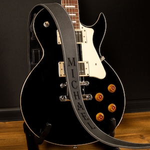 Puede incluir: Una guitarra el&eacute;ctrica negra con perillas doradas y detalles blancos, apoyada en un soporte. Una correa de guitarra gris con el nombre "MICHAEL" en may&uacute;sculas est&aacute; sobre la guitarra. La correa tiene costuras blancas.