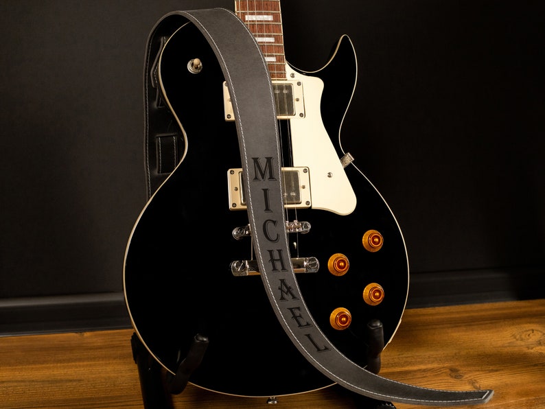Puede incluir: Una guitarra el&eacute;ctrica negra con perillas doradas y detalles blancos, apoyada en un soporte. Una correa de guitarra gris con el nombre "MICHAEL" en may&uacute;sculas est&aacute; sobre la guitarra. La correa tiene costuras blancas.