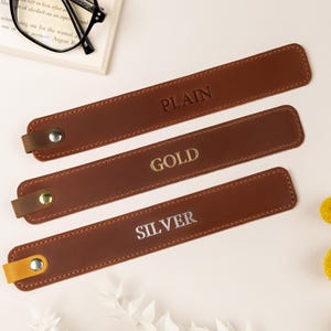 May include: Three brown leather bookmarks with the words "PLAIN", "GOLD", and "SILVER" embossed on them. Each bookmark has a small loop and a metal snap. The bookmarks are arranged on a white surface with a book and glasses in the background.
