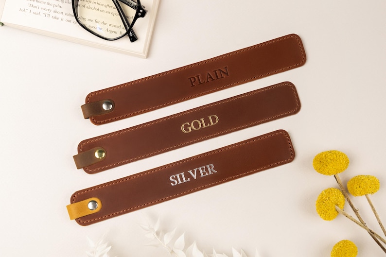 May include: Three brown leather bookmarks with the words "PLAIN", "GOLD", and "SILVER" embossed on them. Each bookmark has a small loop and a metal snap. The bookmarks are arranged on a white surface with a book and glasses in the background.