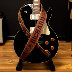 May include: A black electric guitar with a brown leather strap. The strap is embossed with the name "RICHARD" and star designs. The guitar has gold-colored knobs and is on a black stand.