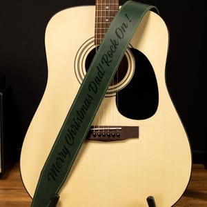May include: An acoustic guitar with a green guitar strap. The strap has the text "Merry Christmas Dad! Rock On!" in black. The guitar is light-colored with a dark sound hole and bridge.