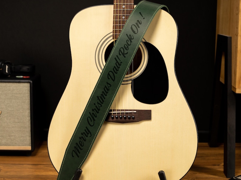 May include: An acoustic guitar with a green guitar strap. The strap has the text "Merry Christmas Dad! Rock On!" in black. The guitar is light-colored with a dark sound hole and bridge.