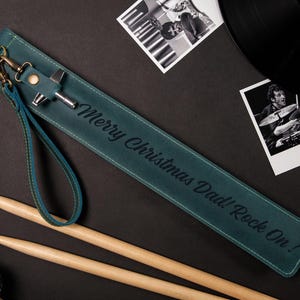 May include: Teal leather drumstick holder with the text "Merry Christmas Dad! Rock On!" and a wrist strap. Includes drumsticks, a microphone, and black and white photos of a drummer and a vinyl record.