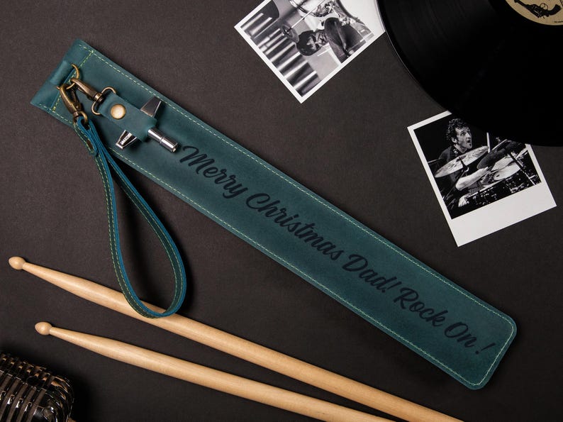 May include: Teal leather drumstick holder with the text "Merry Christmas Dad! Rock On!" and a wrist strap. Includes drumsticks, a microphone, and black and white photos of a drummer and a vinyl record.