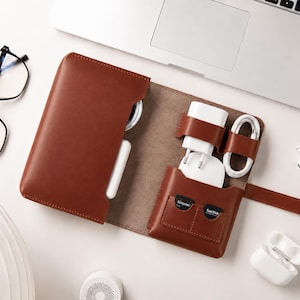 May include: A brown leather tech organiser case featuring multiple interior pockets and elastic loops. It holds a white power adapter, a coiled white charging cable, and two small black memory cards. The case has a snap-button closure strap.