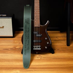 May include: A dark green bass guitar strap with the text "Daddy, we'll always be your biggest fans. Love, Emma, Matt & Theo" and a decorative emblem. A brown bass guitar is in the background.