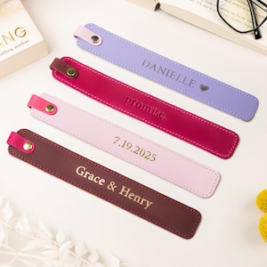 May include: Four personalized leather bookmarks in shades of purple, red, pink, and brown, each with a different inscription in gold lettering. The bookmarks are arranged near a book and decorative elements.