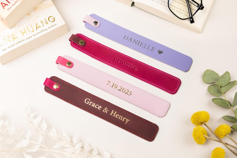 May include: Four personalized leather bookmarks in shades of purple, red, pink, and brown, each with a different inscription in gold lettering. The bookmarks are arranged near a book and decorative elements.