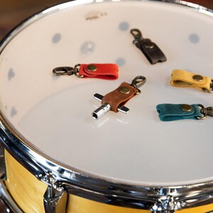 May include: A collection of colorful leather drum keychains in red, brown, yellow, teal, and black, each with a metal clasp. The keychains are displayed on the white surface of a drum.