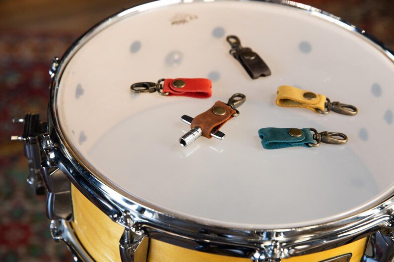 May include: A collection of colorful leather drum keychains in red, brown, yellow, teal, and black, each with a metal clasp. The keychains are displayed on the white surface of a drum.