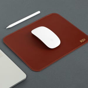 Puede incluir: Un mouse pad de cuero marrón con un ratón de ordenador y un lápiz óptico blancos. El mouse pad tiene un borde cosido e iniciales doradas en la esquina. Un portátil es parcialmente visible en la imagen.