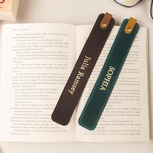 Könnte beinhalten: Zwei Leder-Lesezeichen mit goldfarbenem Schriftzug auf einem offenen Buch. Eines ist dunkelbraun mit "Julia Ramsey", das andere grün mit "Sophia". Beide haben eine Lederschlaufe und einen goldenen Knopf. Eine Brille ist in der Nähe zu sehen.