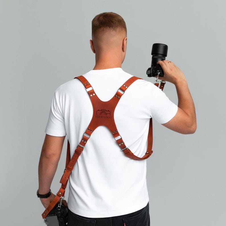 May include: A brown leather camera harness with adjustable straps and metal hardware. The harness features a logo on the back and is designed to hold a camera. The person is wearing a white t-shirt and black jeans.