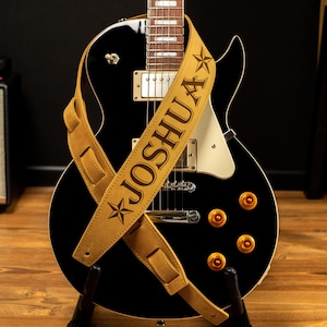 Puede incluir: Una guitarra el&eacute;ctrica negra con una correa de cuero color canela. La correa est&aacute; personalizada con el nombre "JOSHUA" en letras marr&oacute;n oscuro y dise&ntilde;os de estrellas. La guitarra tiene herrajes dorados y est&aacute; sobre un soporte negro.