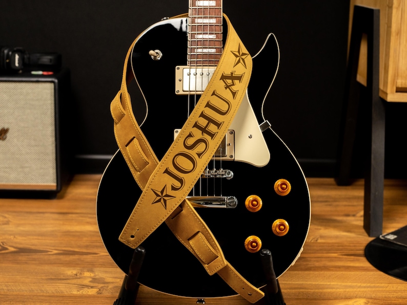 Puede incluir: Una guitarra el&eacute;ctrica negra con una correa de cuero color canela. La correa est&aacute; personalizada con el nombre "JOSHUA" en letras marr&oacute;n oscuro y dise&ntilde;os de estrellas. La guitarra tiene herrajes dorados y est&aacute; sobre un soporte negro.