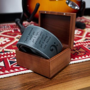 May include: A gray guitar strap with white stitching and the text "Don't practice until you get it right, practice until you can't get it wrong. Love, Melissa" is displayed in a wooden box. A treble clef is also visible.