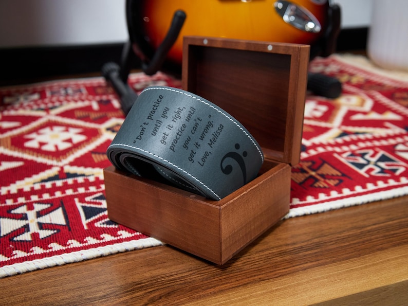 May include: A gray guitar strap with white stitching and the text "Don't practice until you get it right, practice until you can't get it wrong. Love, Melissa" is displayed in a wooden box. A treble clef is also visible.