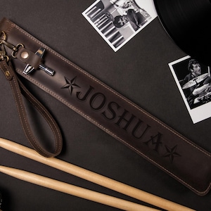 May include: A brown leather drumstick case with the name "JOSHUA" embossed on it, along with star designs. The case has a wrist strap and a metal clasp. Two drumsticks and photos are also visible.