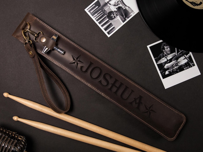May include: A brown leather drumstick case with the name "JOSHUA" embossed on it, along with star designs. The case has a wrist strap and a metal clasp. Two drumsticks and photos are also visible.