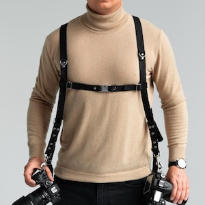May include: Black leather camera harness with adjustable straps and metal hardware. The harness is designed to hold two cameras, with clips at the bottom to attach the cameras. The harness is worn over a beige turtleneck sweater.
