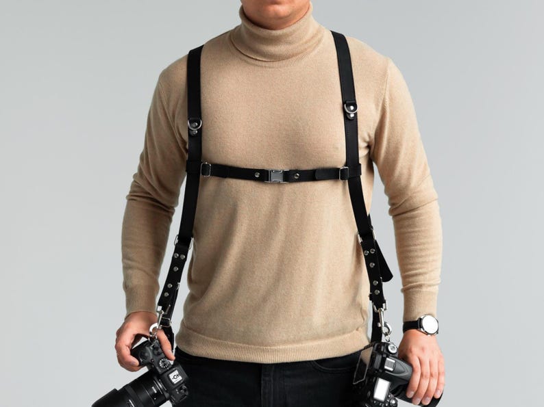 May include: Black leather camera harness with adjustable straps and metal hardware. The harness is designed to hold two cameras, with clips at the bottom to attach the cameras. The harness is worn over a beige turtleneck sweater.