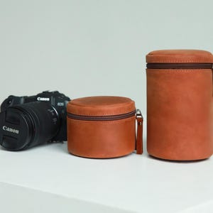 May include: A black Canon EOS camera with a lens cap and two brown leather lens cases. The cases are cylindrical with brown zips and pull tabs. The cases are different sizes, one short and wide, the other tall and narrow.