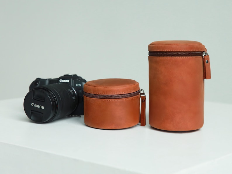 May include: A black Canon EOS camera with a lens cap and two brown leather lens cases. The cases are cylindrical with brown zips and pull tabs. The cases are different sizes, one short and wide, the other tall and narrow.
