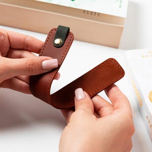 May include: A brown leather bookmark with a dark green accent and a gold-colored snap. The bookmark is shown near a book, suggesting its use as a reading accessory. The leather appears to be stitched for durability.
