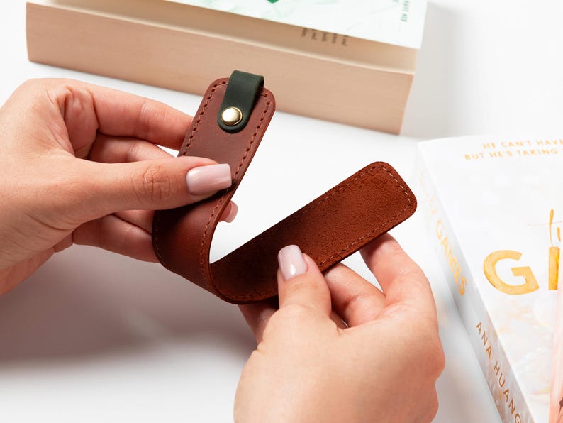 May include: A brown leather bookmark with a dark green accent and a gold-colored snap. The bookmark is shown near a book, suggesting its use as a reading accessory. The leather appears to be stitched for durability.